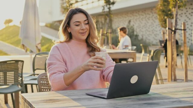 Lovely successful positive caucasian woman, freelancer, small business owner, sits at a table outdoors, drinks coffee during a break at work at a laptop, looks into the distance, thinks, dreams, smile