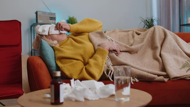 Grandmother ill suffering from cold or allergy pain lying on couch in living room at home. Sick elderly woman with towel on head blows sneezes wipes snot into napkin. Coronavirus quarantine pandemic