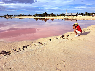 Fototapeta na wymiar Latin adult photographer man with shorts, red shirt and hat takes pictures on the sand next to the pink colored lagoon with a high concentration of salt, Las Coloradas in Yucatan Mexico
