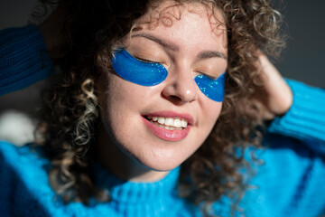 Beautiful smiling young woman with curly hair with eye patches at home, home beauty treatment concept.