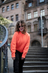 Stylish young woman with curly hair in orange sweater walking in the city