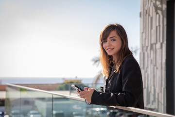 Fototapeta premium A smiling businesswoman answers emails on her smartphone from her office balcony