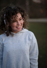Young curly haired woman in grey sporty suit in headphones has fun outdoors in the evening