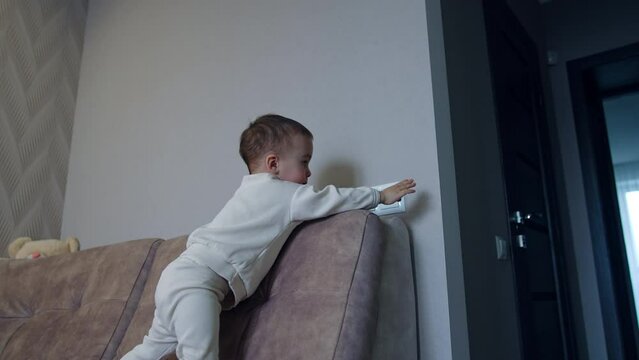 Active kid walks by the sofa and reaches the light switch. Baby switches on illumination in the room. Low angle view.