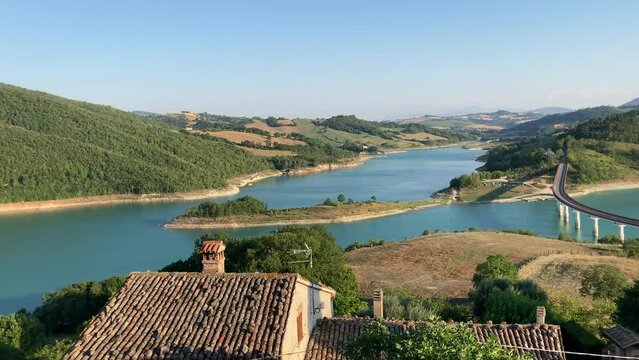 Wide-sweeping view from Castreccioni castle ruins at Cingoli Lake (Lago di Castreccioni) with glowing mirroring azure waters, its tall bridge, chiesa di San Giorgio and the Sibillini suffused in haze