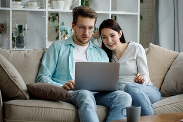 A young man and a woman are sitting on the sofa with a laptop and laughing.