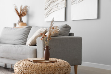 Vase with dry reeds, flowers and books on rattan pouf in living room