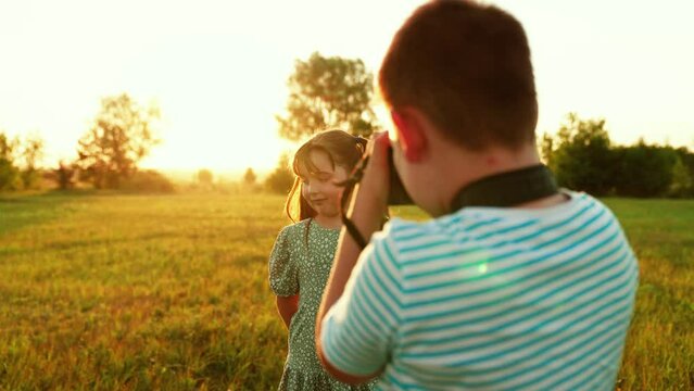 Hobby of children is taking pictures. Child girl poses as model for boy to photographer with camera. Children are photographed in park at sunset with digital camera. Childrens games in nature in sun.