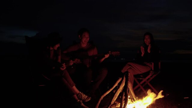 Asian friends camping holiday, Sitting around a bonfire, drinking beer, playing guitar in the dusk, Lifestyle Relaxation, Vacations Camping trip, Travel