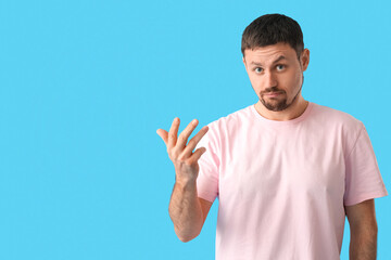 Fototapeta premium Confused young man in pink t-shirt gesturing with hand, expressing uncertainty and questioning on blue background, his facial expression shows doubt and questioning
