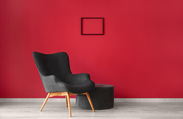 Stylish grey armchair, pouf and frame near red wall