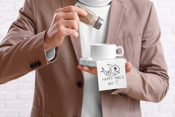 Young man sprinkling pepper in cup of coffee on white brick background, closeup. April Fools' Day prank - obrazy, fototapety, plakaty