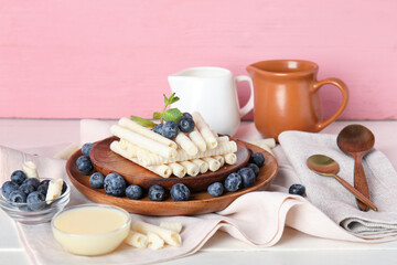 Plates with delicious wafer rolls and blueberries on white wooden table against pink background