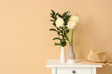 Vases with ranunculus flowers, plant branches and hat on table near beige wall