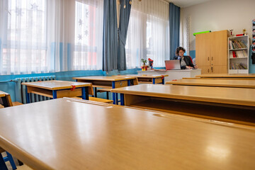 Female teacher sitting and writing notes in her notebook in empty classroom. Lots of empty desk in front of her. Horizontal indoors shot with copy space. Back to school concept.