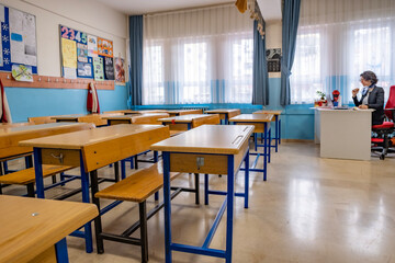 Empty class room with wood tables and female teacher sitting at her desk, reading notes in her notebook. Horizontal indoors shot with copy space. Back to school concept.