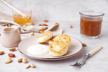 Plate with tasty cottage cheese pancakes with sour cream, caramel syrup and nuts on light background