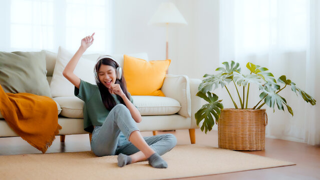 Young asian woman listening to music on on the floor beside couch in living room at home. Happy asian beautiful female relax dancing and wearing headset
