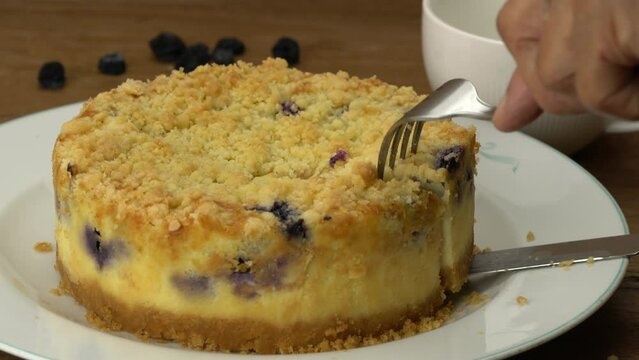 Using knife and fork taking a slice of delicious homemade freshly baked bluberry crumble cheesecake in white ceramic plate on wooden table.