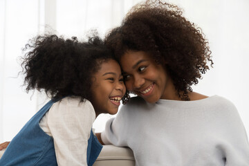 Happy family embracing together on sofa at home. Smiling African American young mother spending time with her child daughter at home
