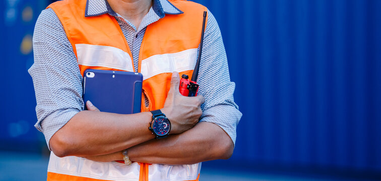 Crop image, man engineer arm crossed in uniform arm crossed holding walkie talkie and tablet standing containers yard. Area logistics import export and shipping.