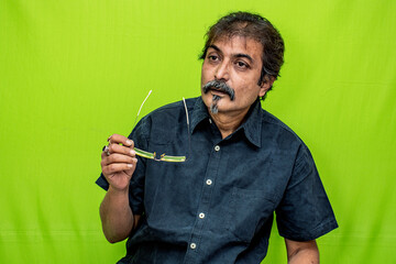 A well-dressed Indian man wearing a black shirt is standing against a green screen, looking calm and pensive while holding a spectacle in his right hand
