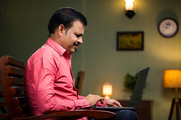 Middle aged man busy working on laptop on chair at home - concept of technology, entrepreneur and surfing internet.