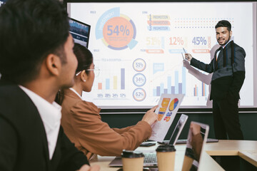 A cheerful and confident Asian businessman stands, presenting bar charts data from the projector...