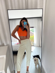 Fit tanned woman in orange top and cosy comfy pants at home take photo selfie on phone in mirror for social media, stories, vertical.