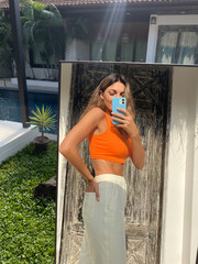 Fit tanned woman in orange top and cosy comfy pants at home outdoor take photo selfie on phone in mirror for social media, stories, vertical.