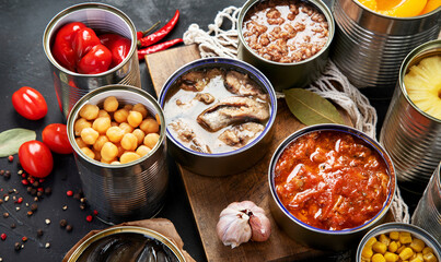 Canned vegetables, beans, fish and fruits in tin cans on black background. Food stocks.