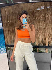 Fit tanned woman in orange top and cosy comfy pants at home outdoor take photo selfie on phone in...