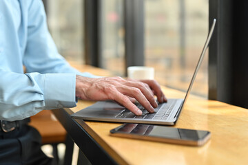 Cropped image of man employee sitting by office window, using laptop computer on wooden counter