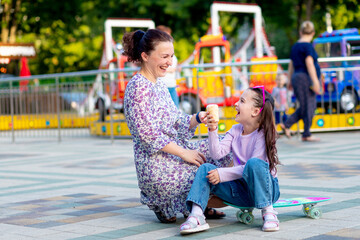 a child girl with her mother in an amusement park in the summer eating ice cream near the...