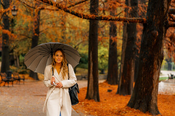 Woman with umbrella taking a walk in autumn park on rainy day, orange and red leaves have fallen on the ground