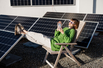 Young woman talks on phone while sitting relaxed on rooftop with a solar station. Happy owner of...