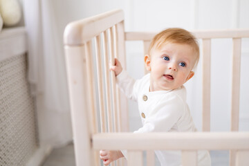 close-up of a smiling baby 6 months old blond boy in a crib in a bright bedroom in a white cotton bodysuit, portrait, concept of children's goods