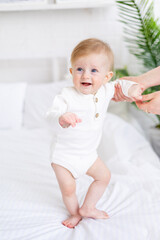 mom's hands are holding a baby 6 months old, a blond boy with blue eyes on a white bed in a light bedroom in a white cotton bodysuit, the concept of children's goods