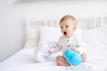 yawning baby 6 months old blond boy with a toy in his hands on a white bed in a bright bedroom in a cotton bodysuit wants to sleep