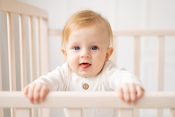 portrait of a 6-month-old blond boy standing in a crib in a bright children's room and smiling in a cotton bodysuit, the concept of children's goods