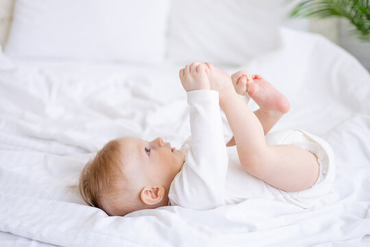 a 6-month-old blond boy plays with his legs on a white bed in a bright bedroom after sleeping in the morning in a cotton bodysuit, the concept of children's goods