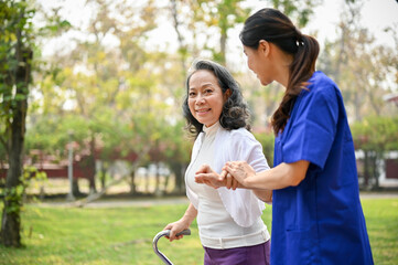 Happy old Asian disabled lady with a walking stick being helped by a caring female caregiver