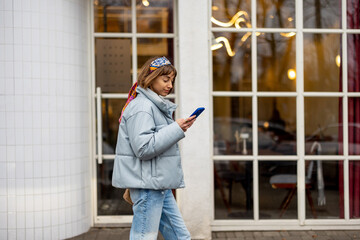 Young stylish woman in blue casual clothes walks with phone on background of modern cafe window...