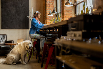 Woman sits with her cute adorable dog at modern coffee shop and works on laptop. Pet friendly...