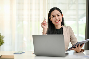 Charming Asian businesswoman sits at her desk with tablet in her hand, smiling and looking at camera.