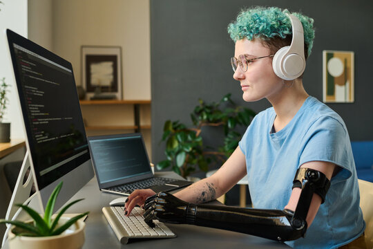 Young developer in wireless headphones typing security codes on computer sitting at her workplace at home