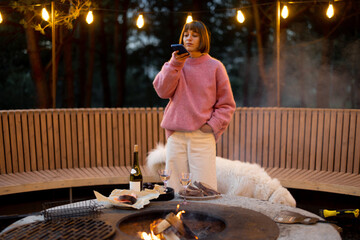 Fototapeta premium Young woman spends leisure time with her dog, taking photo on phone at beautiful lounge area with bonfire and round bench in pine forest at dusk