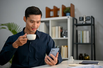 Happy Asian businessman using his smartphone while sipping morning coffee in the office.
