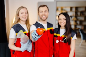 A group of young medical people in uniform stand in the middle of the classroom and smile after a first aid lesson. A man holds a red heart in his hands. Concept of assistance