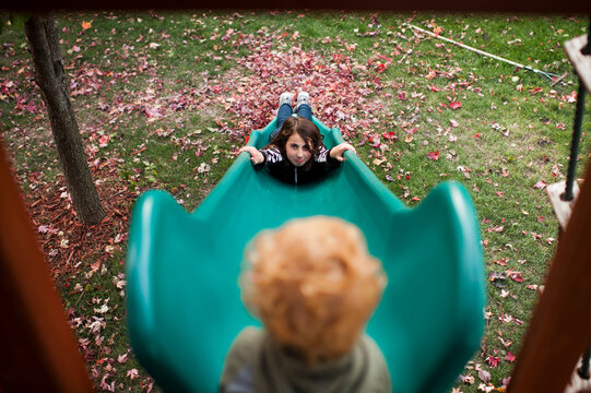 Girl slides down slide while looking up at her brother with pouty face